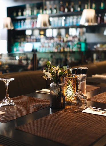Ambiance de Restaurant Romantique : Table Dressée et Bar Table de restaurant élégante avec fleurs blanches, bougie et verres à vin devant un bar flou.